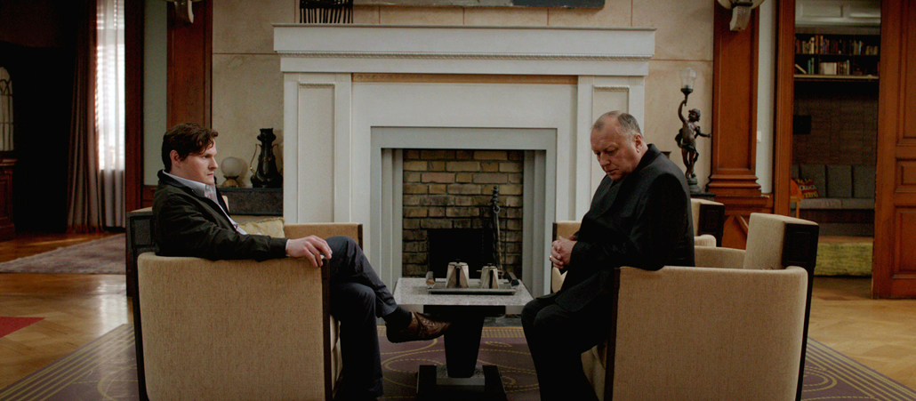 Der Mörder und der Bischof sitzen sich in zwei Sesseln gegenüber. Während Oliver den Bischof anstarrt, schaut dieser nachdenklich zu Boden.