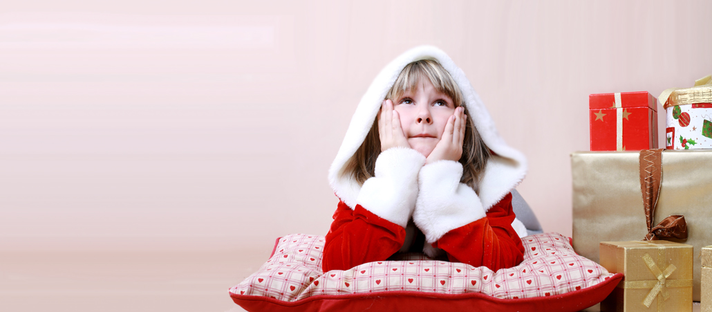Mädchen im Weihnachtsmantel stützt sich auf ein Kissen und wartet, daneben Geschenke