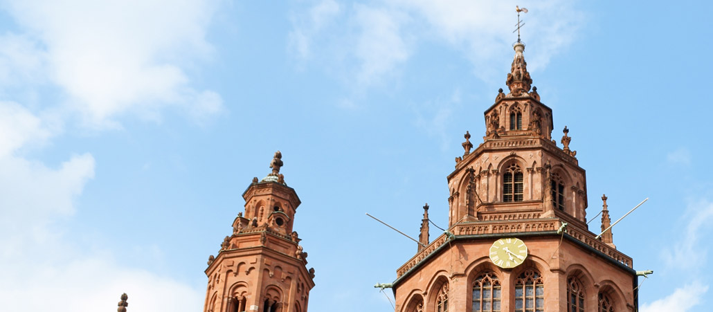 Blick von unten auf den Turm des rötlichen Mainzer Doms, blauer Himmel im Hintergrund
