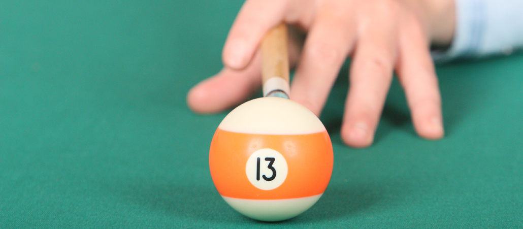 Eine Billiardkugel mit der Nummer 13; hinter ihr eine Hand und ein Stock kurz vor dem Stoß.