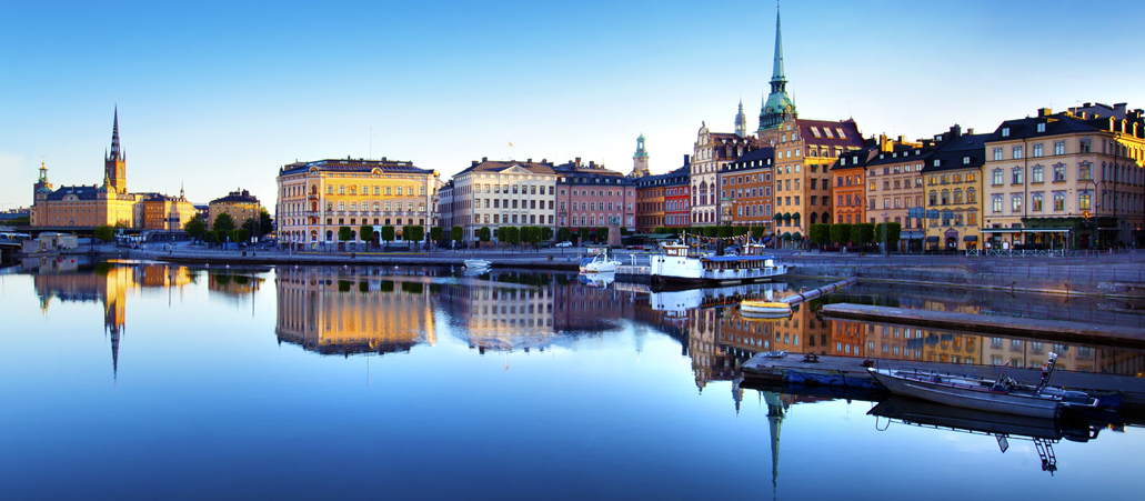 Die bunten Häuser der Altstadt von Stockolm heben sich vom blauen Himmel ab und spiegeln sich im Wasser.