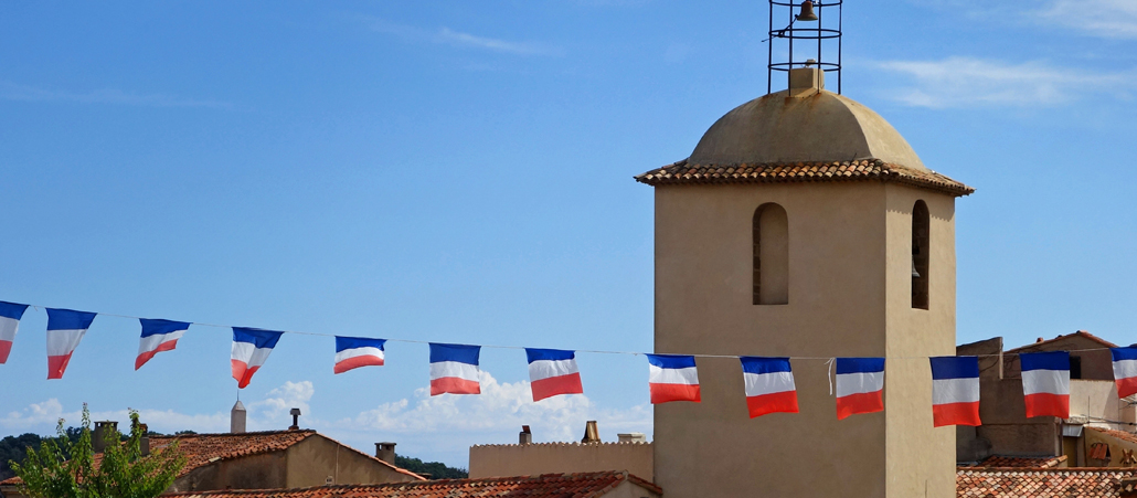 KIrchturm, Fahnenkette der Französichen Fahne von links nachs rechts durch das Bild, vor blauem Himmel, Häuserdächer
