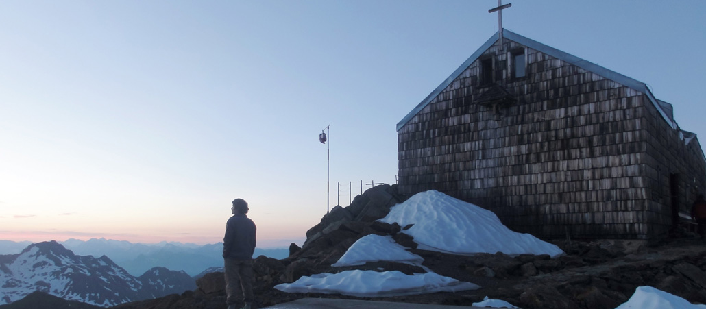 Eine holzvertäfelte Kapelle in den Bergen, davor steht ein Besucher, der ins Tal schaut.