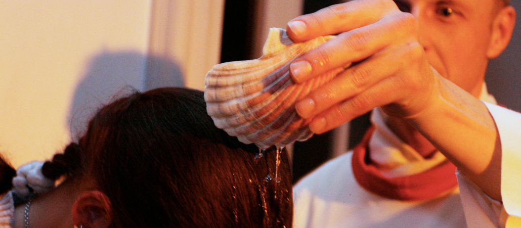 Ein Priester gießt Wasser aus einer Muschel über den geneigten Kopf einer Frau.