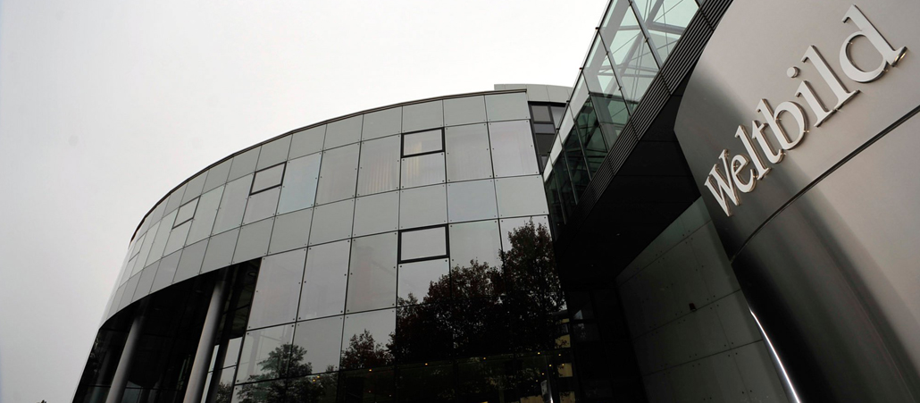 Ein rundes Glashaus vor einem wolkenverhangenen Himmel. Am rechten Bildrand ist das Firmenlogo mit der Aufschrift 