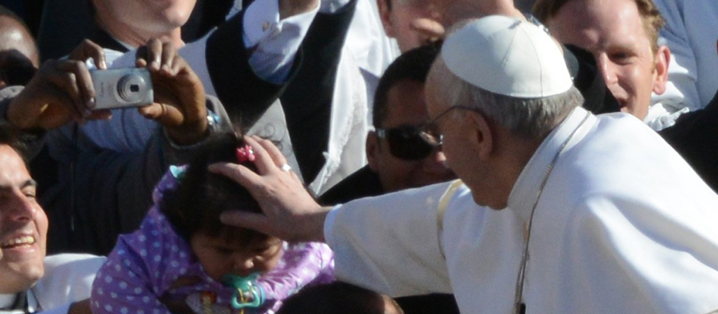 Papst Franziskus reckt die Hand nach einem Kind, welches ihm hingehalten wird, und segnet es.