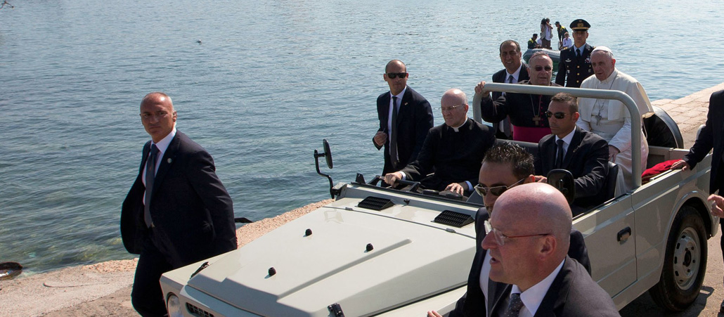 Papst Franziskus sitzt hinten auf einem Geländewagen. Sechs Sicherheitskräfte gehen neben dem Auto her. Im Hintergrund sieht man das Meer am Hafen der Mittelmeerinsel Lampedusa.