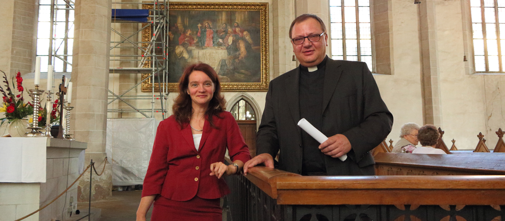 Eine zierliche Frau im roten Kleid und ein stämmiger Mann stehen locker an ein Gitter gelehnt im Kirchenschiff.