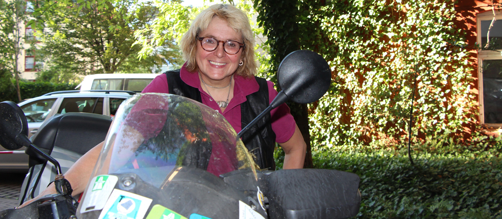 Eine blonde Frau steht in einem lilanen Polohemd und einer scharzen Weste neben ihrem Motorrad (und wird von der Frontscheibe halb verdeckt).