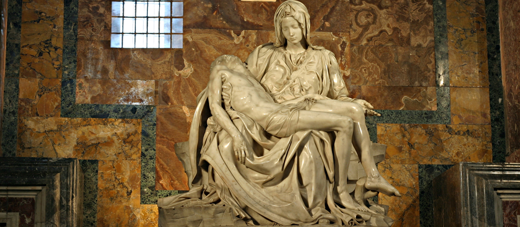 Zwei aus einem Marmorblock gefertigte Figuren: Die jung wirkende Maria hält ihren toten Sohn Jesus in den Armen. Im Hintergrund ist die dunkle Marmorverkleidung des Petersdoms und ein kleines Fenster.