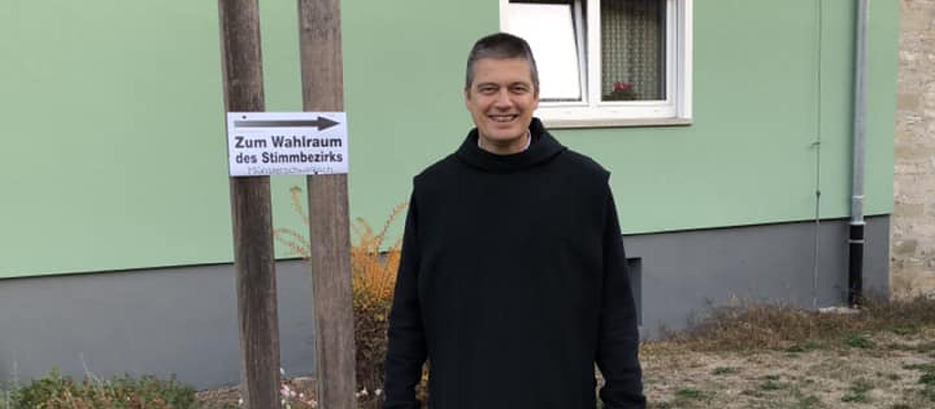 Pater Christoph vor einem Wegweiser zum Wahllokal bei der bayerischen Landtagswahl 2018