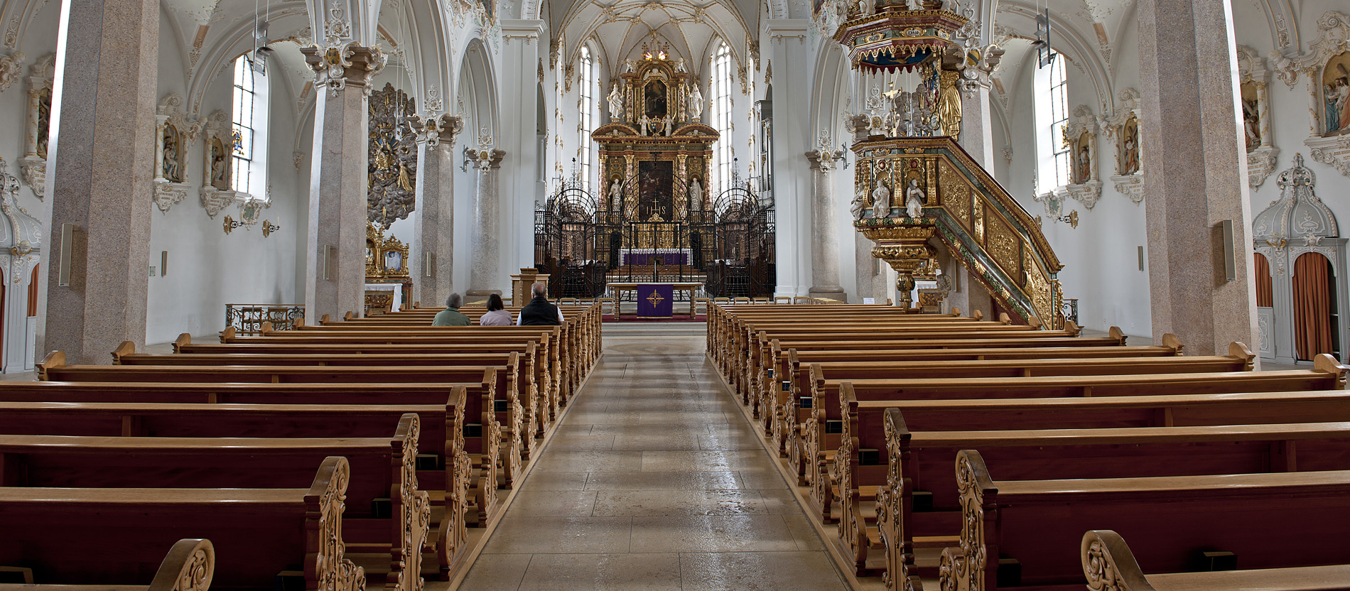 Blick in das fast vollständig leere Schiff der Klosterkirche in Mariastein, Schweiz. Nur drei Menschen sitzen in den Bänken.