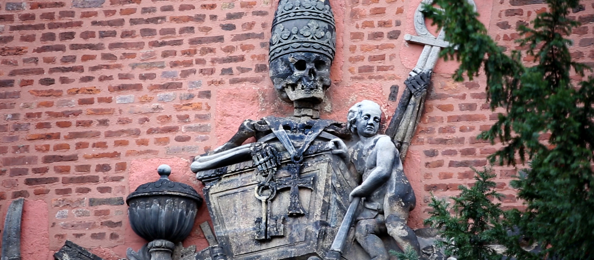 Ein Relief auf einer Backsteinmauer zeigt ein Skelett in einem Sarg, das eine Papstkrone trägt.