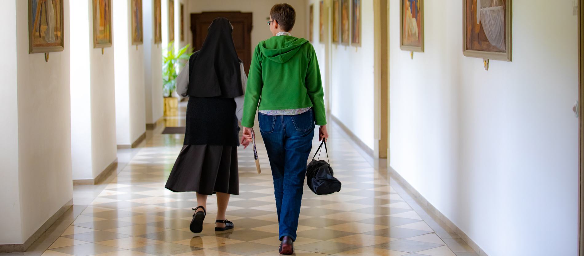 Eine Frau trägt eine Tasche und einen Ordner und geht zusammen mit einer Ordensschwester einen Gang entlang
