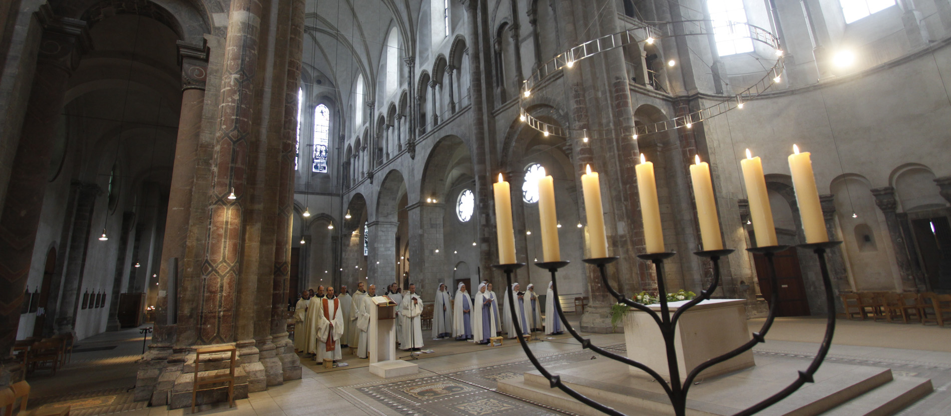 Blick durch die Arme eines siebenarmigen Leuchters auf den Altarraum der Kirche Groß Sankt Martin in Köln, weiter hinten stehen Männer und Frauen in Ordenskleidung