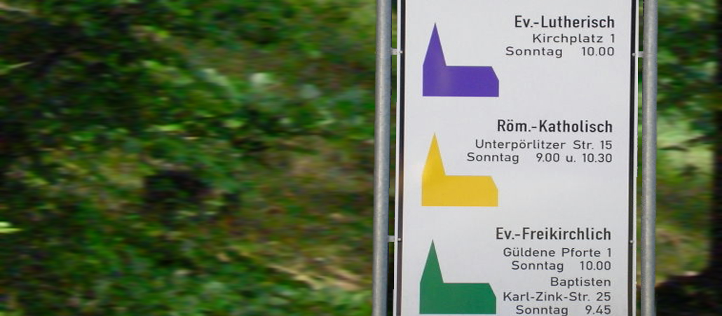 Ein Schild zeigt drei stilisierte Kirchen und die Uhrzeiten der Gottesdienste an.