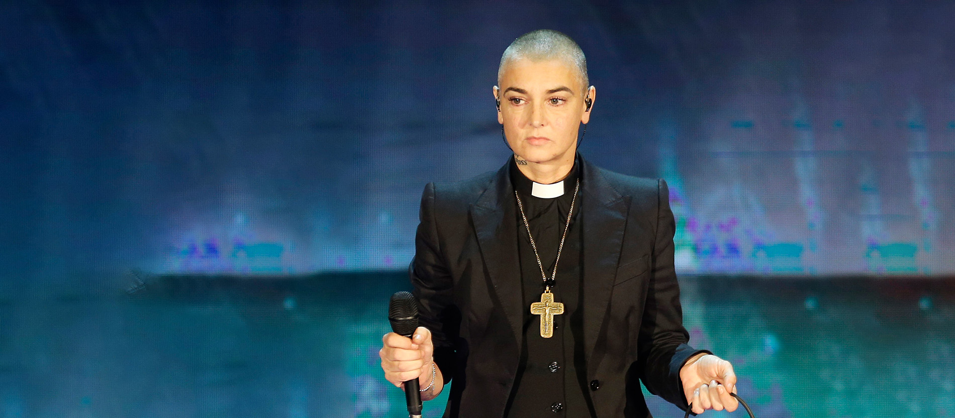 Sinead O'Connor steht mit Kollarhemd, schwarzem Anzug und Kreuz auf einer Bühne