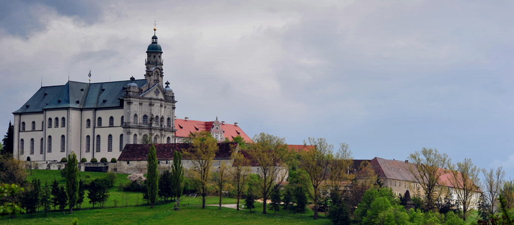 Links im Bild eine weiße, barocke Kirche, unten im Bild mehrere Gebäude eines Klosters. Dunkle Wolken und Herbstwetter.