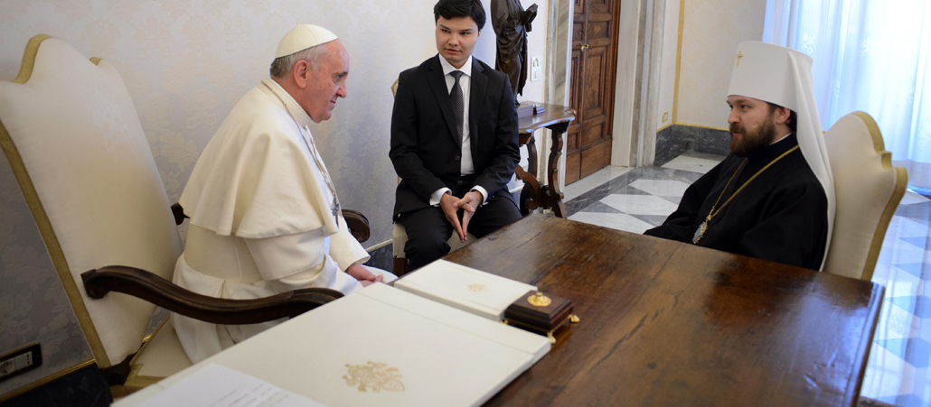 Papst Franziskus sistzt links, hinter seinem Schreibtisch im Vatikan, am rechten Bildrand sitzt der Metropolit Hilarion. Zwischen den beiden sitzt ein Übersetzer.
