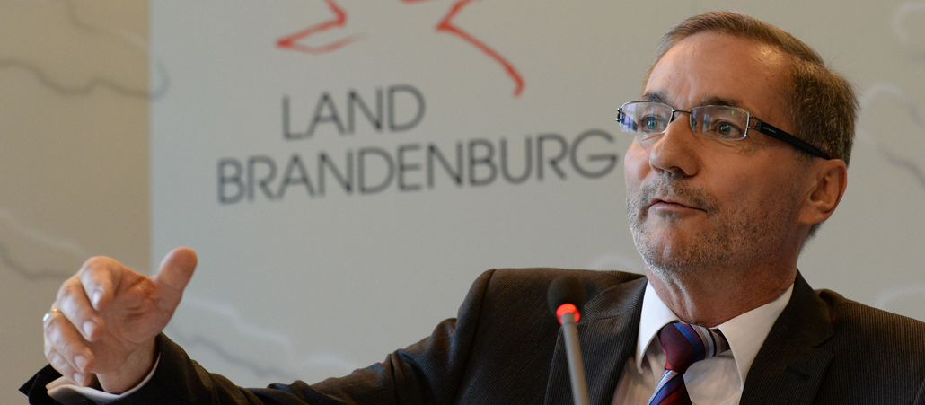 Matthias Platzeck sitzt im schwarzen Anzug vor einer weißen Wand und gestikuliert auslandend mit der rechten Hand.