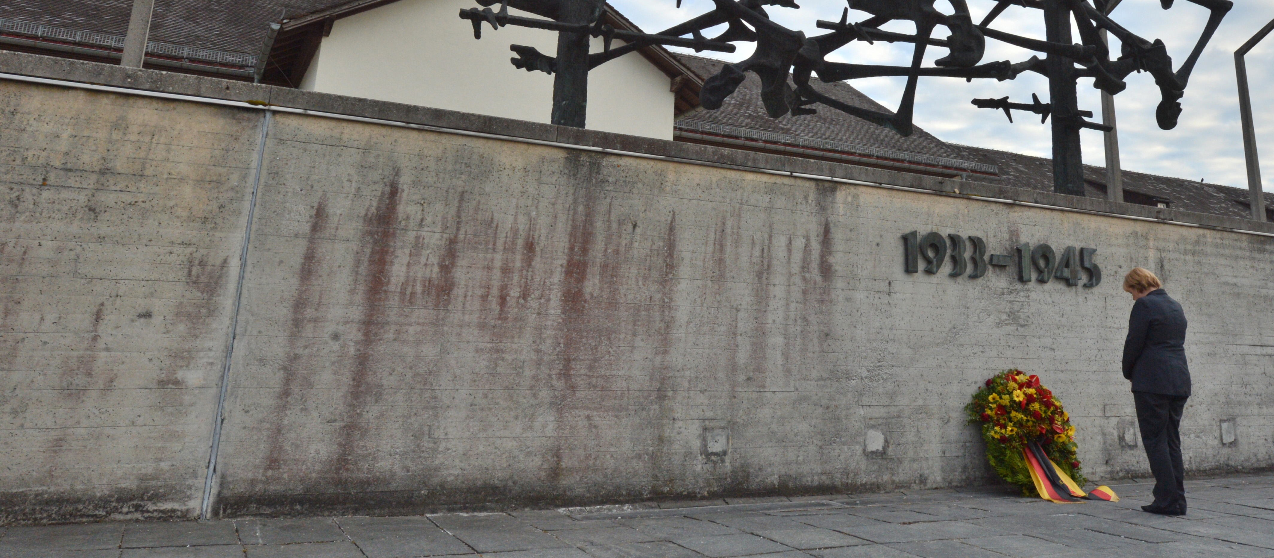 Angela Merkel steht mit gesenktem Kopf vor einer grauen Betonwand mit der Aufschrift 1033-1945. Vor der Wand lehnt ein Kranz.
