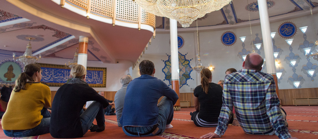 Mehrere Menschen sitzen in einem großen Raum auf dem Teppichboden.