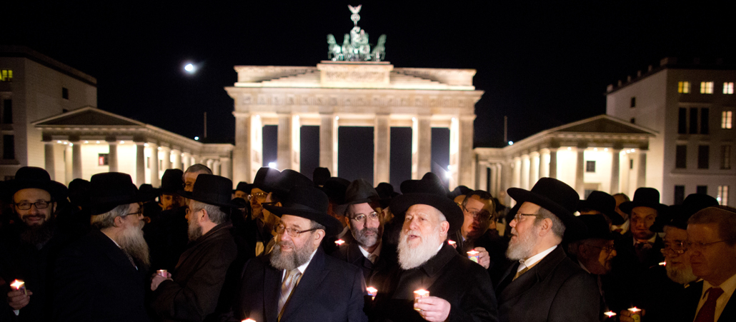 Mehrere schwarz gekleidete Rabbis mit langen Bärten stehen nachts mit Kerzen in der Hand vor dem erleuchteten Brandenburger Tor.