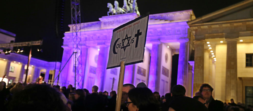 Eine Frau hält vor dem Branenburger Tor eine Fahne mit dem Halbmond, Davidstern und Kreuz hoch.
