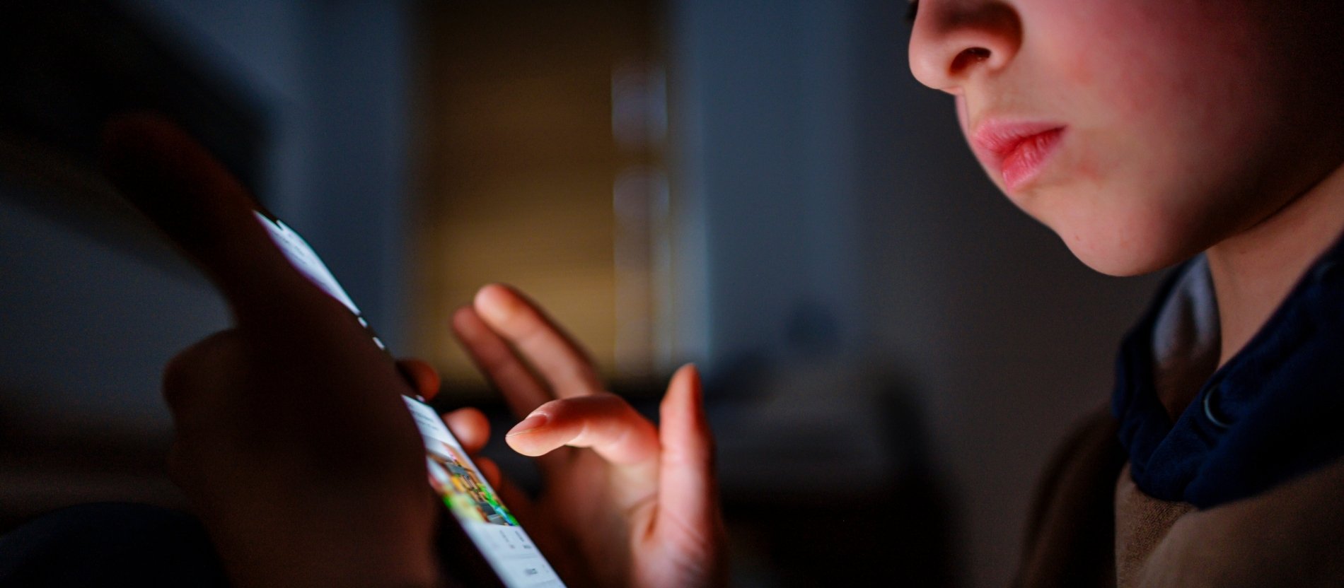 Ein Junge sitzt in einem dunklen Raum und wird von einem Smartphone beleuchtet.