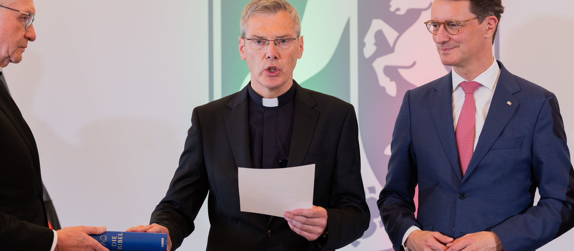 Heiner Wilmer (M.), ernannter Bischof von Münster, legt bei seiner Vereidigung in der Staatskanzlei neben Hendrik Wüst (r., CDU), Ministerpräsident von Nordrhein-Westfalen, seine Hand auf eine Bibel.
