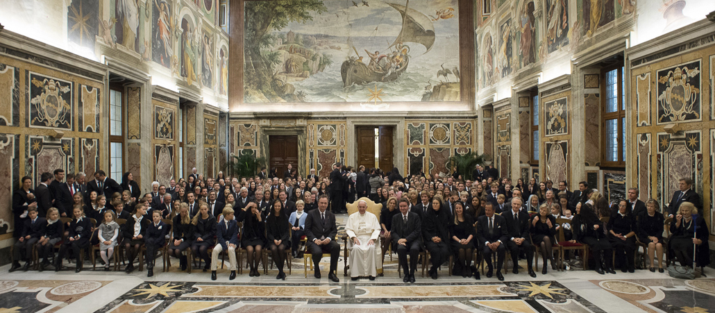 Hunderte festlich gekleidete Leute von jung bis alt sitzen in einem festlichen Saal. Papst Franziskus sitzt in der Mitte.