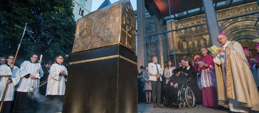 Ein Bischof steht vor einem goldenen Haus, das auf eine Stele angebracht ist.