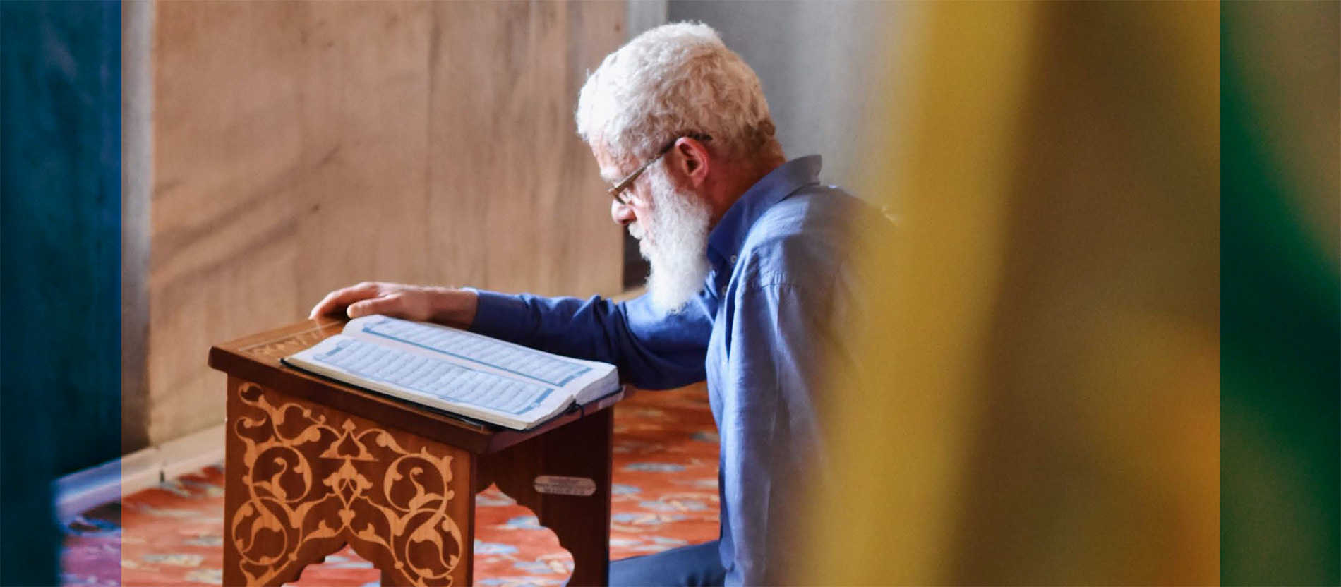 Ein Mann mit weißem Bart sitzt in einer Moschee auf dem Teppichboden und liest den Koran.