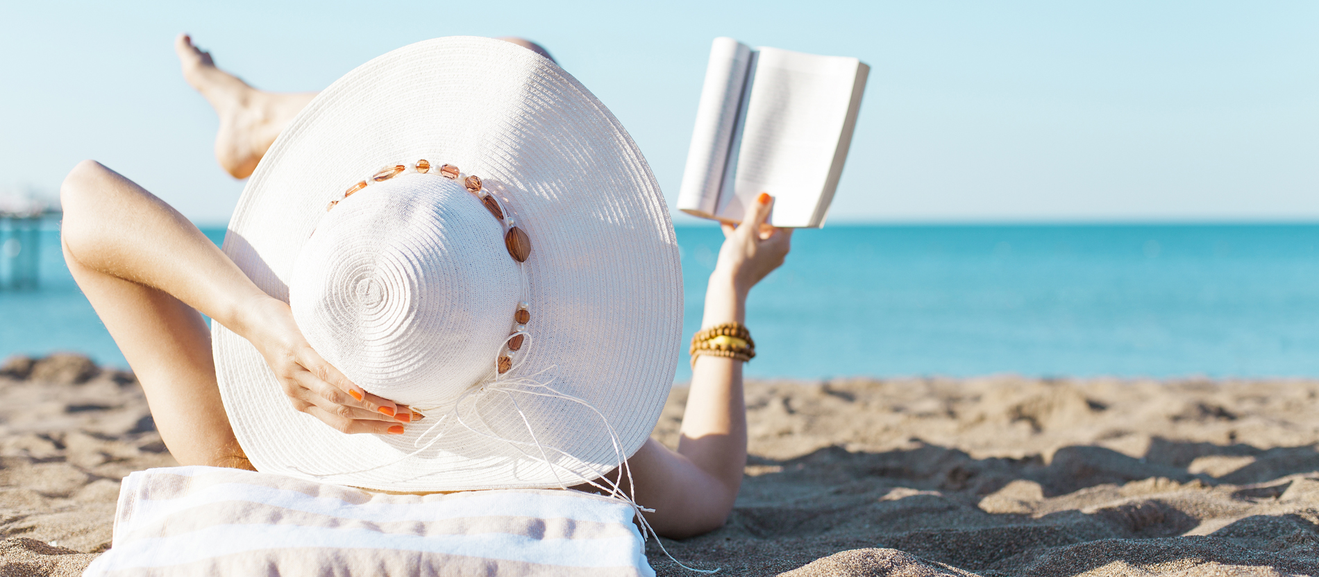 Eine Frau mit einem großen Sonnenhut liegt am Strand und liest in einem Buch.