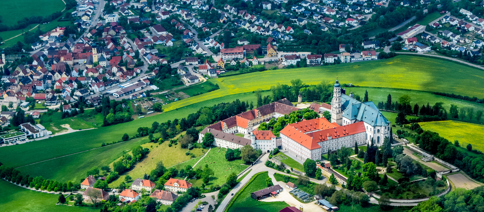Aus der Luft sieht man ein großes Klostergelände mit Kirche auf einem Hügel. Zu Füßen des Hügels sieht man in der oberen Bildhälfte die Stadt Neresheim.