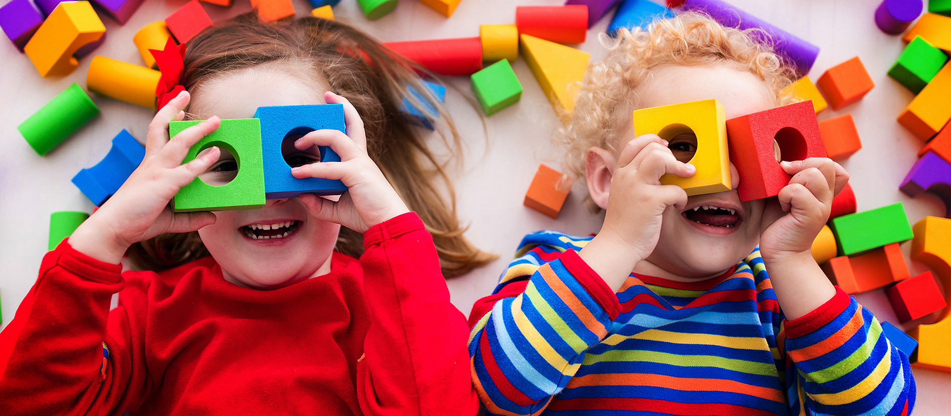 Zwei Kinder liegen rücklings auf dem Boden und spielen mit bunten Bauklötzen. Beide schauen durch je zwei Klötze mit Loch, die sie sich wie eine Brille vor die Augen gelegt haben.