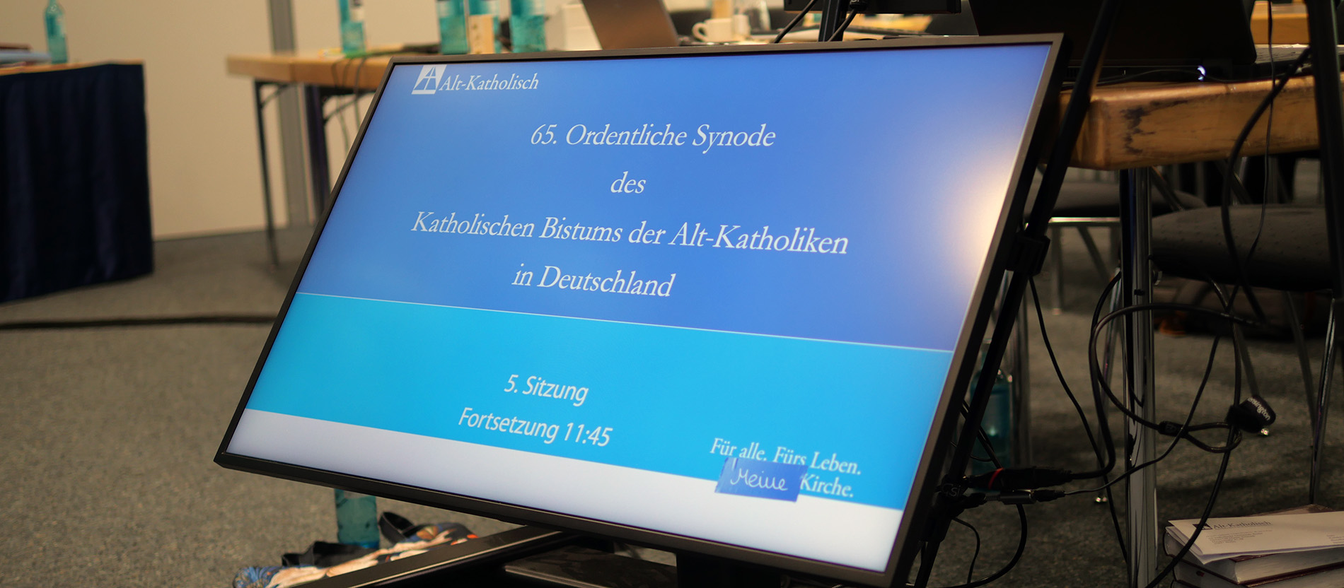 Ein Bildschirm während der alt-katholischen Synode 2026 steht vor dem Tisch des Präsidiums