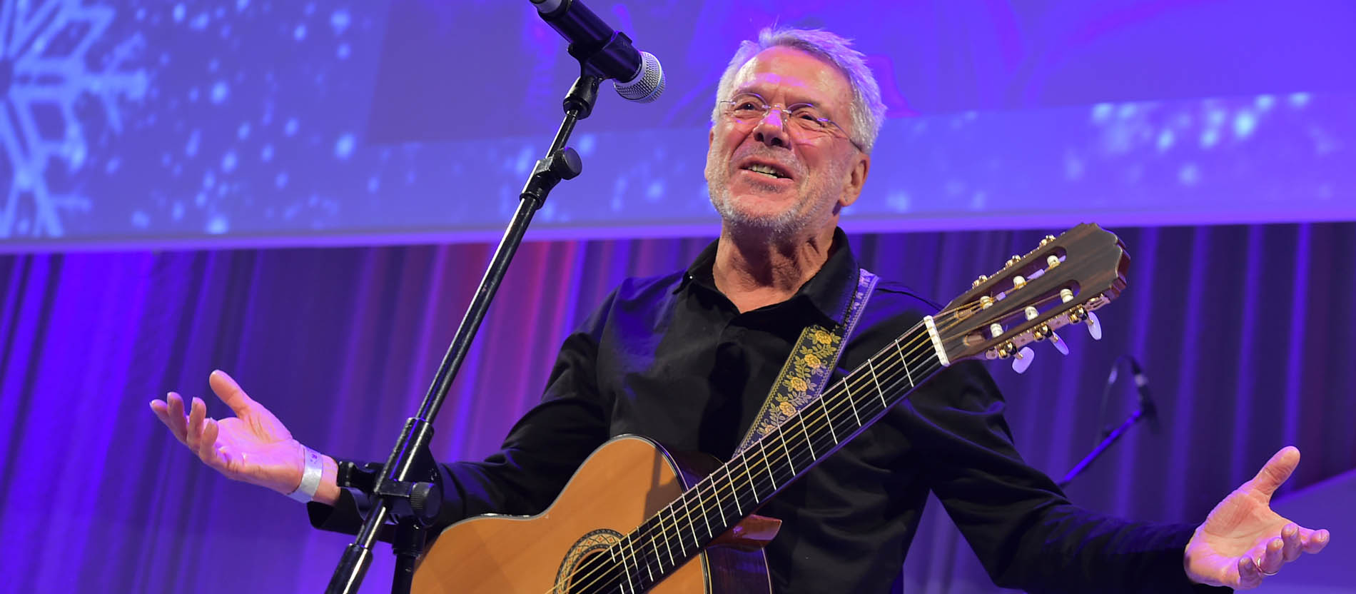 Der Liedermacher Reinhard Mey steht mit Gitarre vor einem Mikrofon und lächelt mit ausgebreiteten Armen.