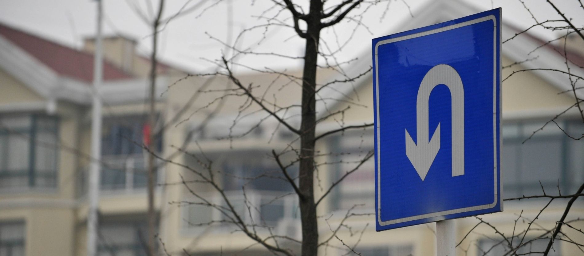 Ein weißer Pfeil auf einem blauen Schild zeigt einen U-Turn an, im Hintergrund kahle Bäume und Häuser