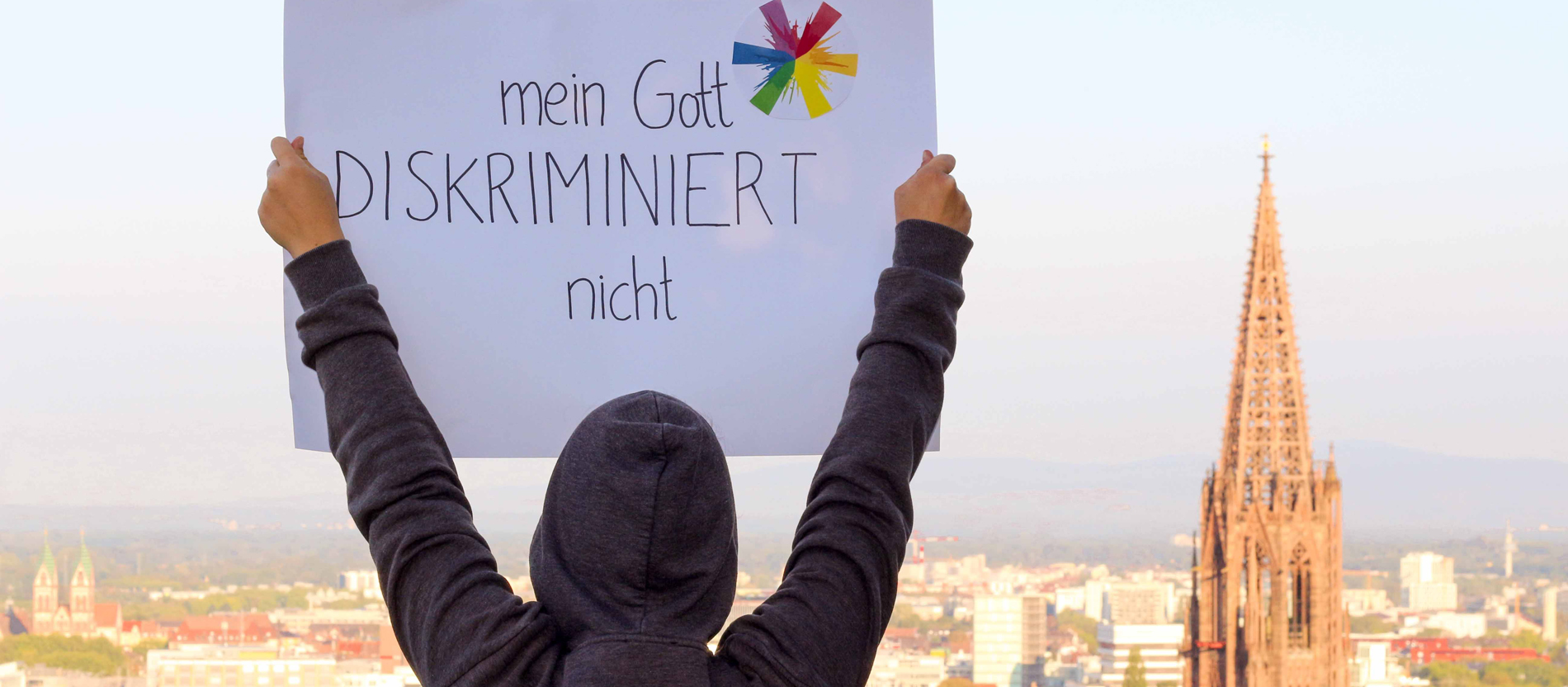 Ein Mensch mit Kapuzenpullover trägt ein Schild mit der Aufschrift "Mein Gott diskriminiert nicht"