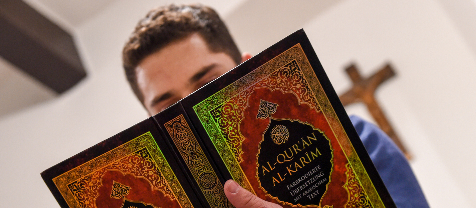 Ein junger Muslim liest im Koran, im Hintergrund sieht man ein Kreuz