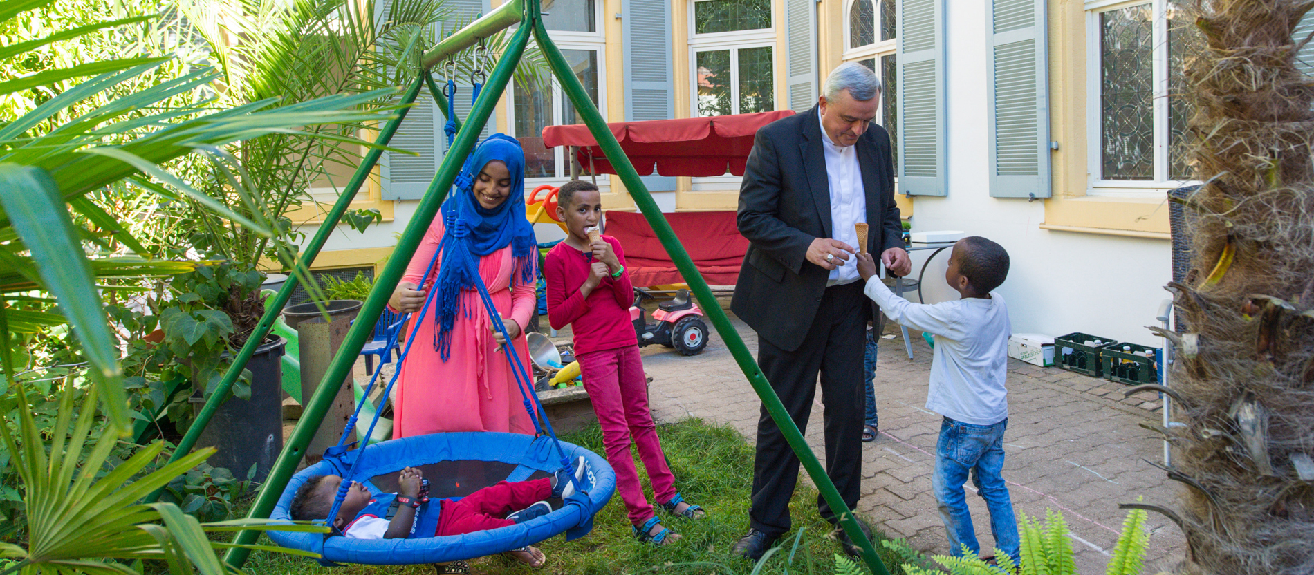 Karl-Heinz Wiesemann spielt im Garten des Vikarienhofs mit den Kindern der somalischen Flüchtlingsfamilie. Die Jungen essen Eis, die Mutter schaukelt einen ihrer Söhne in einer Schaukel.