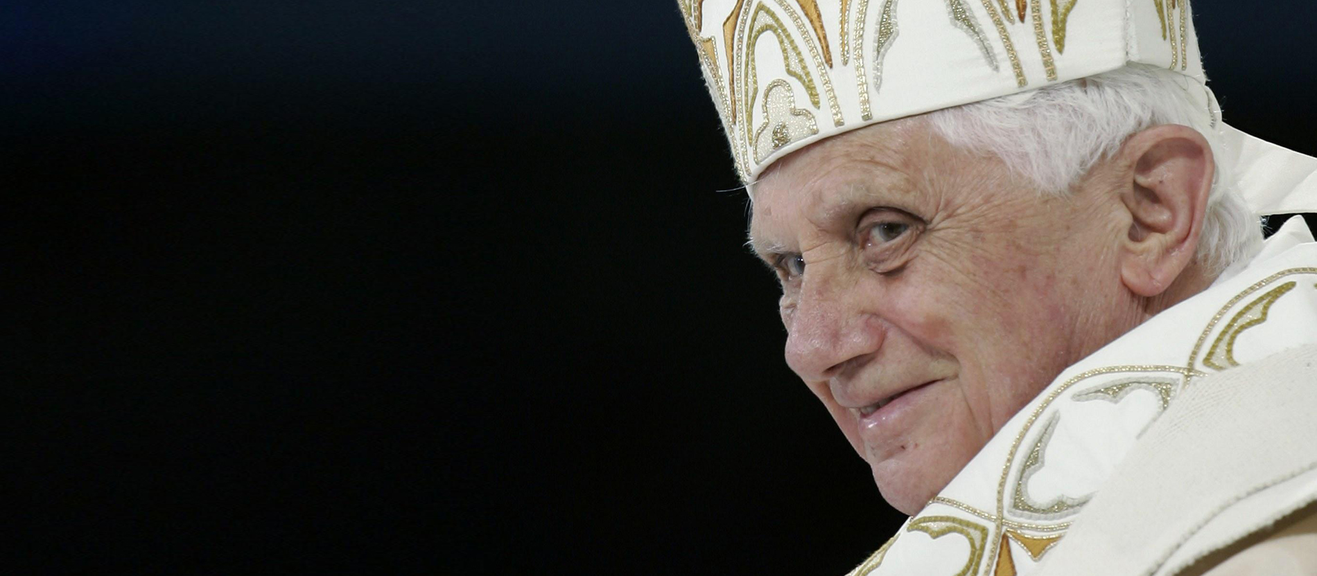 schwarzer Hintergrund, am linken Bildrand das Gesicht des lächelnden Benedikt XVI.