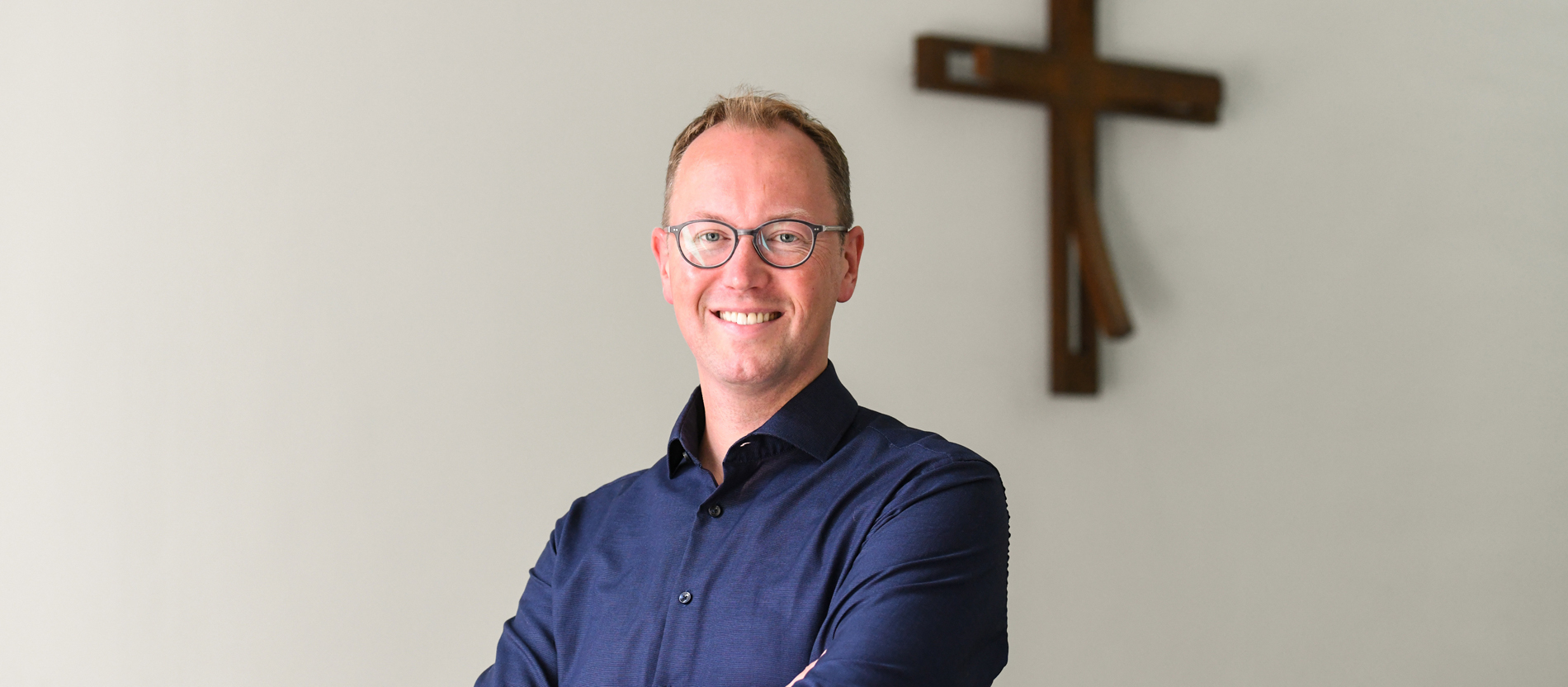 Der evangelische Theologe Christoph Urban steht vor einer weißen Wand, an der ein Kreuz hängt.