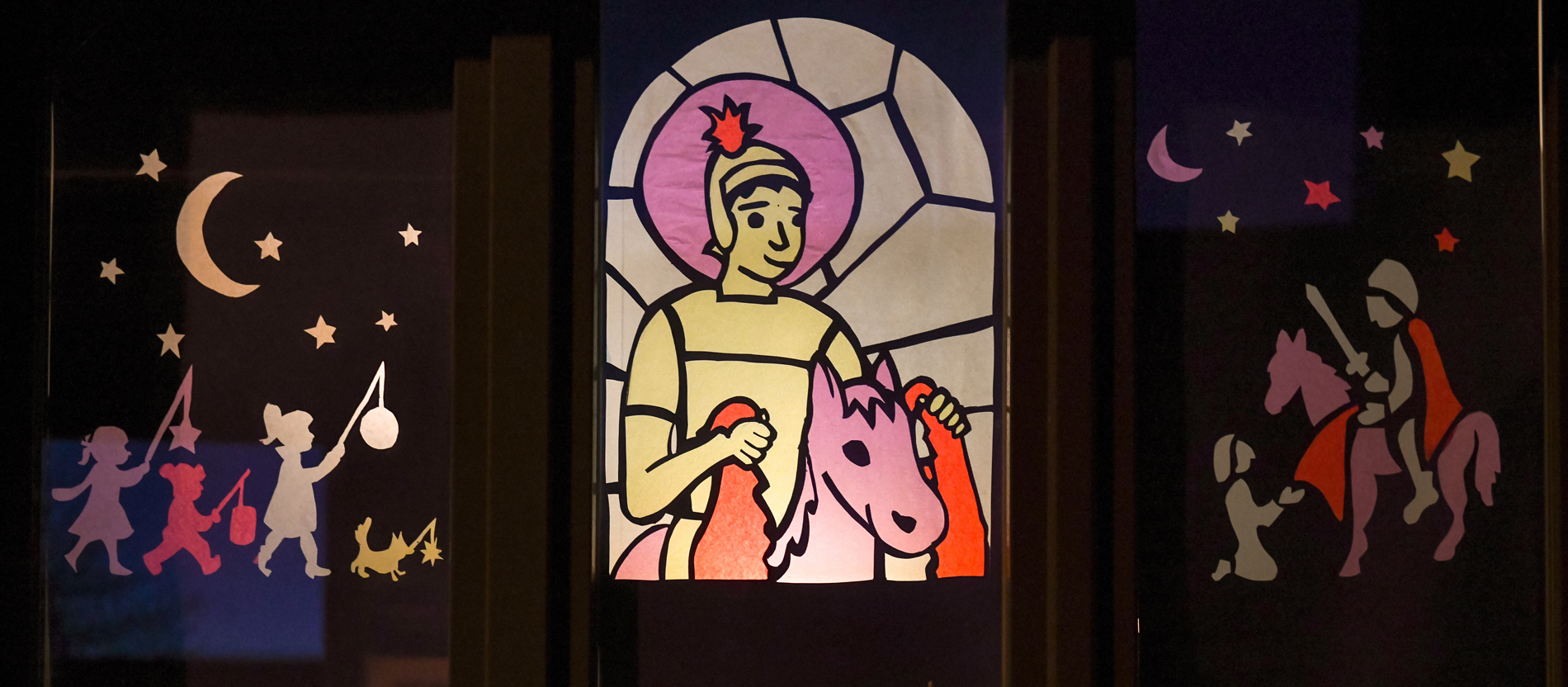 Ein Fensterbild zeigt einen Martinszug mit Kindern mit Laternen und den heiligen Sankt Martin auf dem Pferd