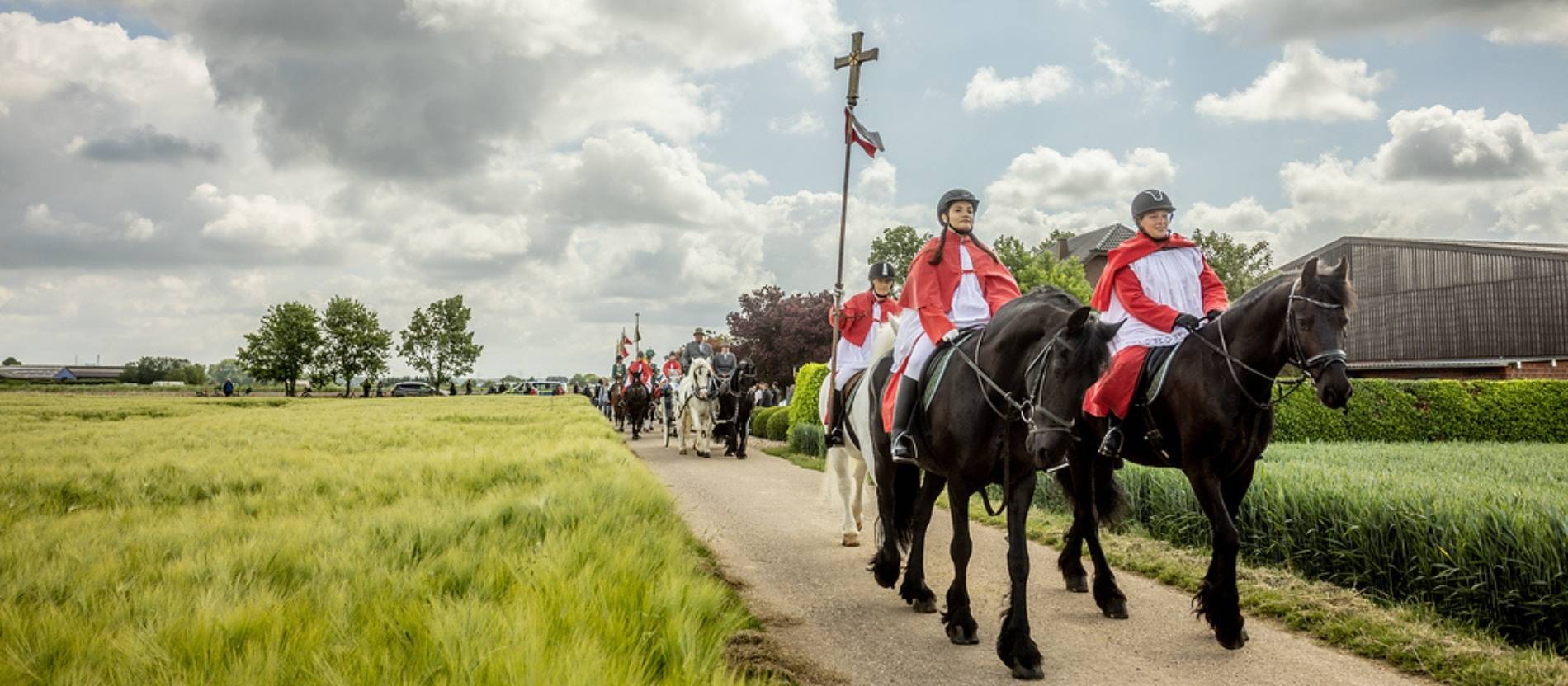 Reiterinnen in Ministrantenkleidung, eine trägt das Vortragekreuz, reiten auf Pferden über ein Feld bei der Reiterprozession.