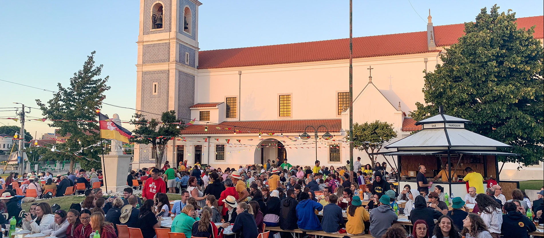 Menschen aus verschiedenen Ländern sitzen an Tischen bei einem Gemeindefest unter freiem Himmel vor einer Kirche in Oia (Portugal).