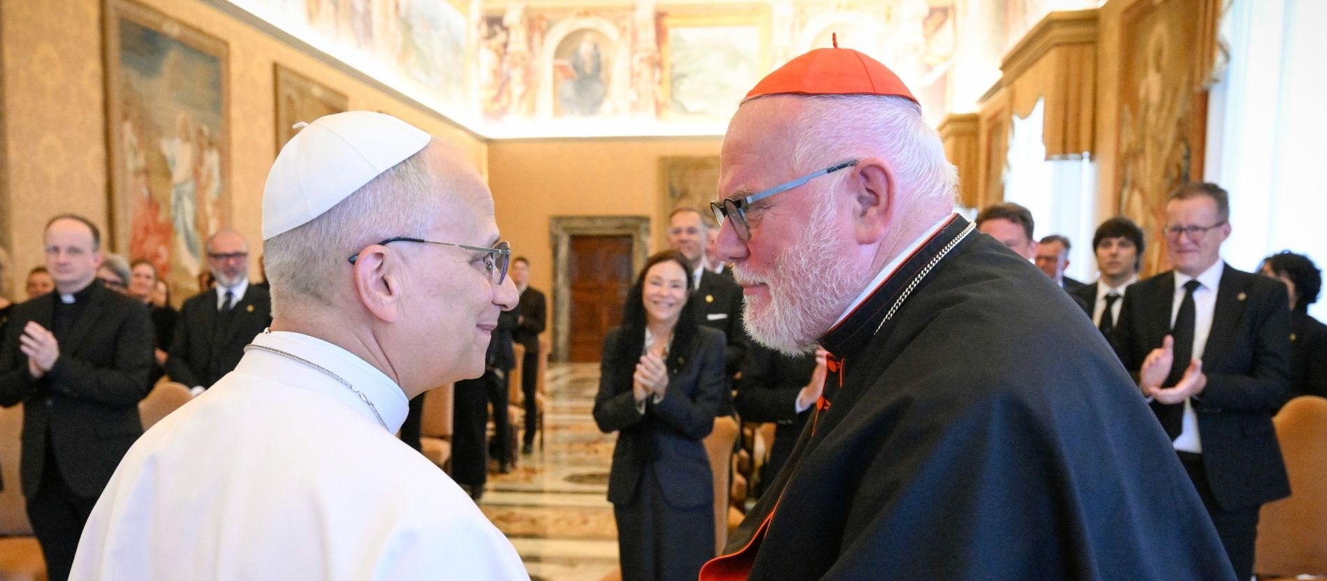 Papst Leo XIV. und Kardinal Reinhard Marx schütteln sich die Hände, im Hintergrund applaudieren Gäste