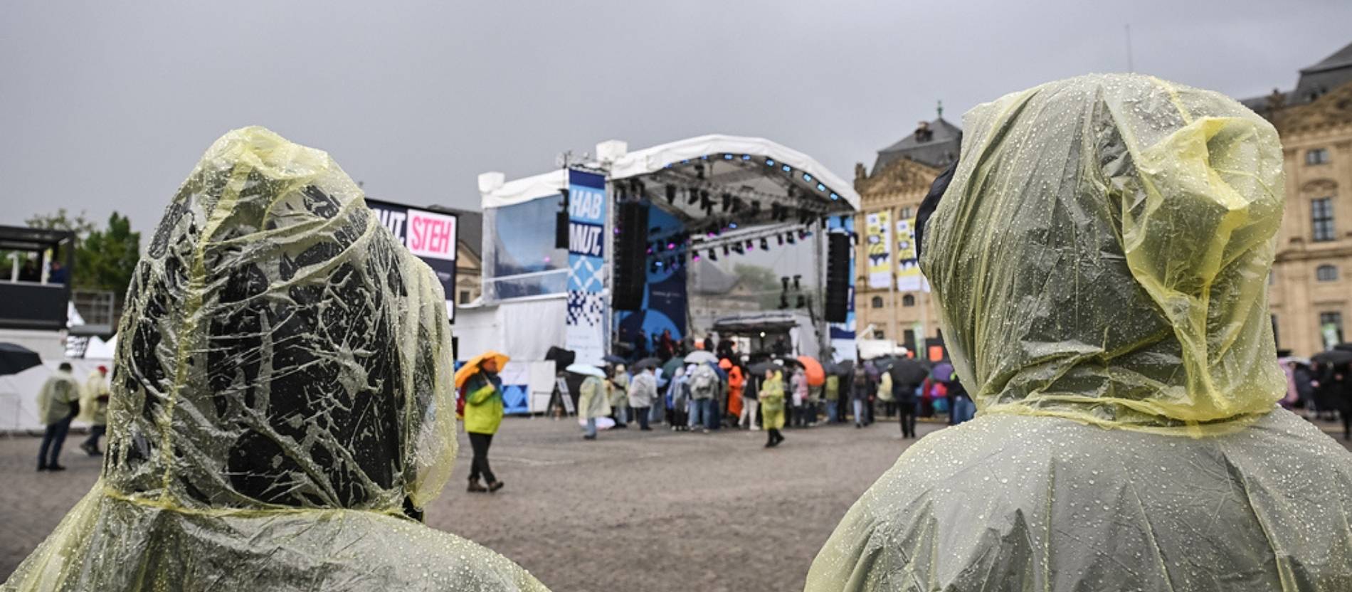 Besucher in Regenponchos, im Hintergrund eine Bühne auf dem Residenzplatz, vor der Eröffnung des 104. Katholikentags in Würzburg.