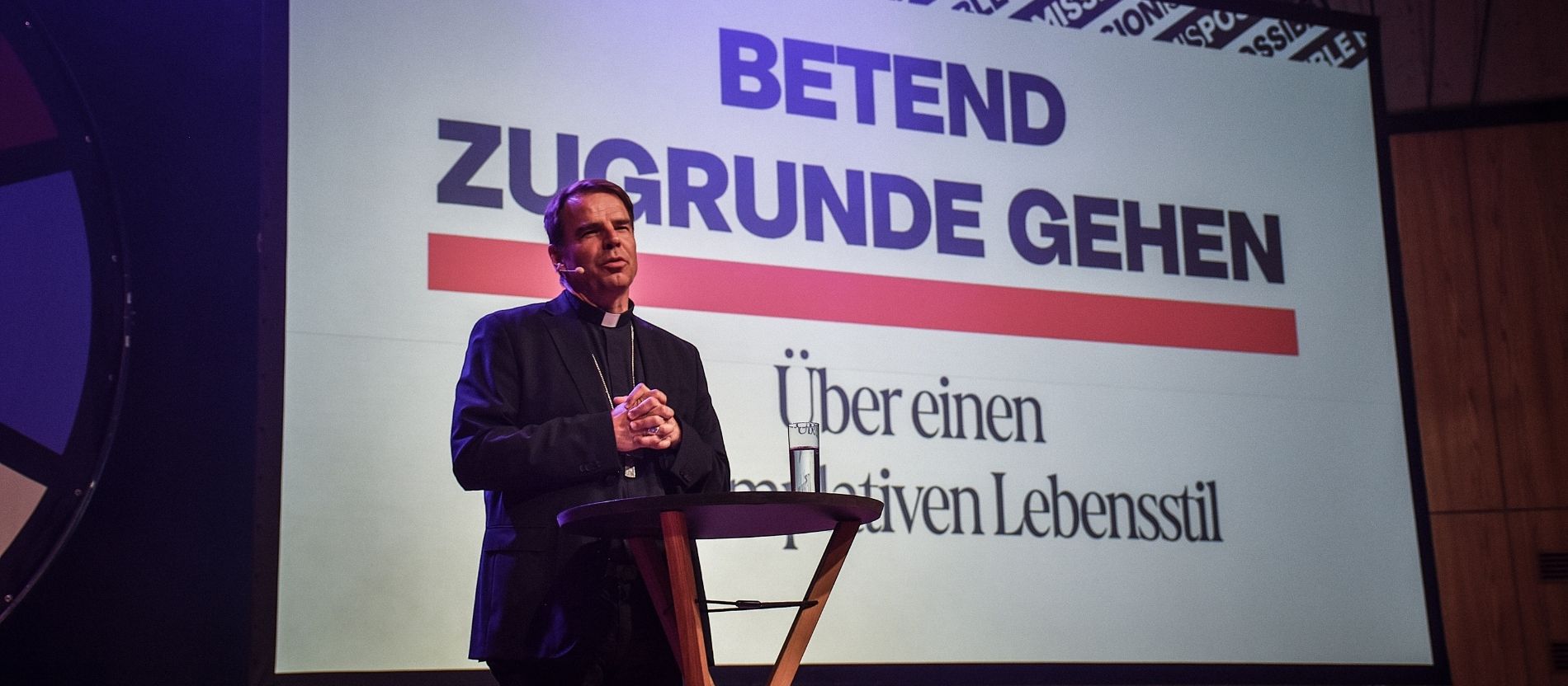 Bischof Stefan Oster spricht beim Missionskongress, im Hintergrund eine Leinwand mit der Aufschrift "Betend zugrunde gehen"